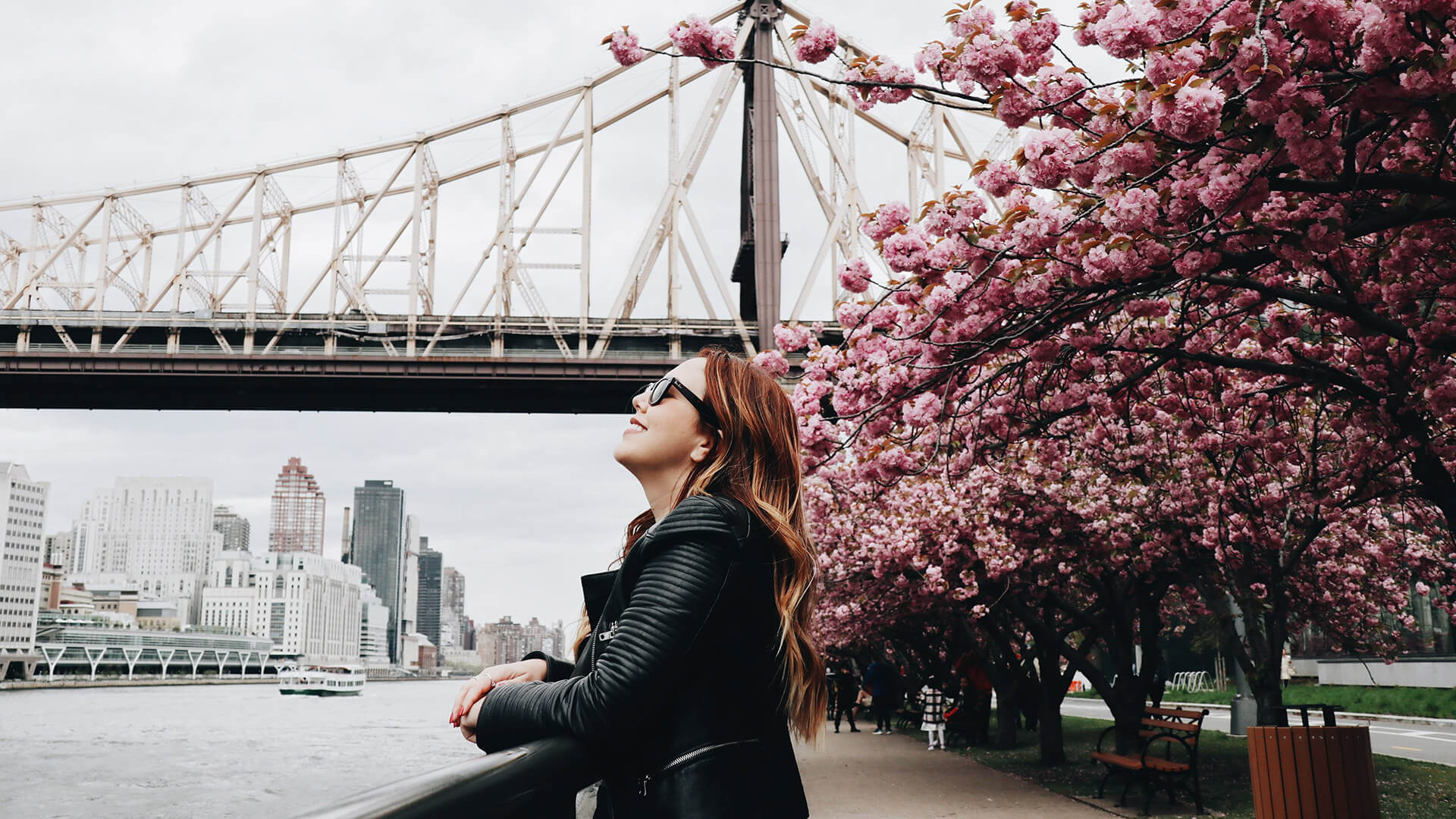 Red haired woman in sunglasses and jacket in LIC next to bridge and cherry blossom smiling and looking up at sky.